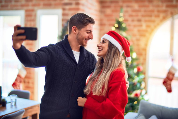 Νεαρό όμορφο ζευγάρι χαμογελά ευτυχισμένη και αυτοπεποίθηση. Στέκεται και αγκαλιάζει κάνει selfie με κάμερα γύρω από το χριστουγεννιάτικο δέντρο στο σπίτι - Φωτογραφία, εικόνα