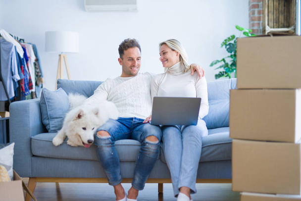 Jeune beau couple avec chien assis sur le canapé en utilisant un ordinateur portable à la nouvelle maison autour des boîtes en carton
 - Photo, image