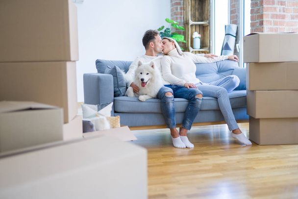 Joven hermosa pareja con perro besándose sentado en el sofá en el nuevo hogar alrededor de cajas de cartón
 - Foto, imagen