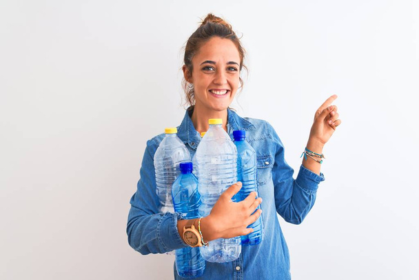 Young redhead woman holding plastic bottle to recycle over isolated background very happy pointing with hand and finger to the side - Foto, Imagen