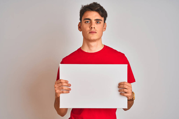 Teenager αγόρι κρατώντας διαφημιστικό πανό με κενό χώρο πάνω από απομονωμένο φόντο με μια σίγουρη έκφραση στο έξυπνο πρόσωπο σκέψης σοβαρή - Φωτογραφία, εικόνα