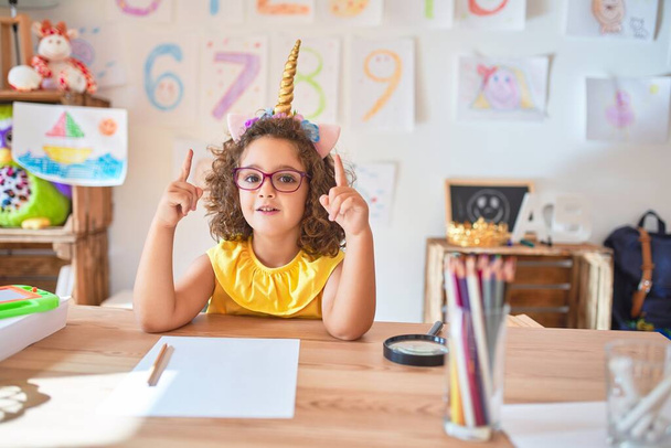 Beautiful toddler wearing glasses and unicorn diadem sitting on desk at kindergarten amazed and surprised looking up and pointing with fingers and raised arms. - Valokuva, kuva