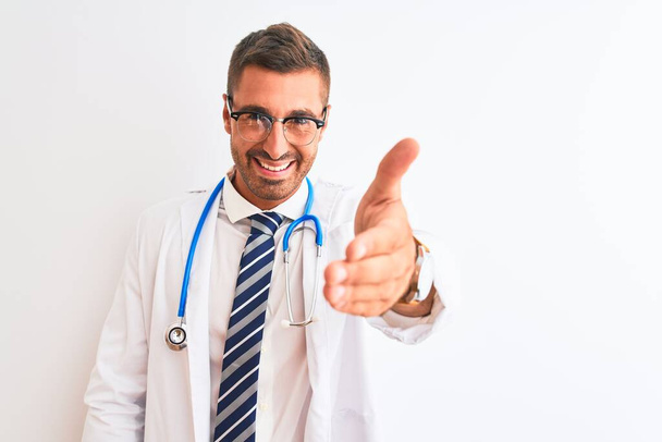 Молодой красивый врач, носящий стетоскоп на изолированном фоне, улыбаясь, дружелюбно предлагая рукопожатие в качестве приветствия и приветствия. Успешный бизнес
. - Фото, изображение