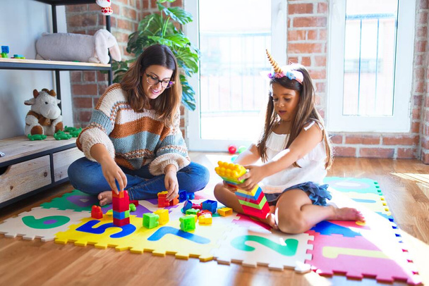 Belle enseignante et tout-petit jouant avec des blocs de construction jouet autour de beaucoup de jouets à la maternelle
 - Photo, image