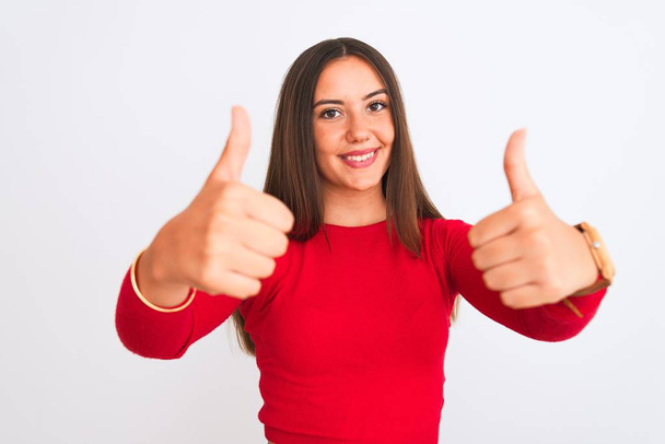 Молодая красивая девушка в красной футболке, стоящей на изолированном белом фоне, одобряя позитивный жест рукой, улыбается и радуется успеху. Жест победителя
. - Фото, изображение