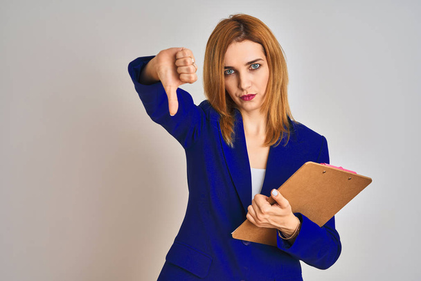 Redhead business caucasian woman holding clipboard over isolated background with angry face, negative sign showing dislike with thumbs down, rejection concept - Foto, afbeelding
