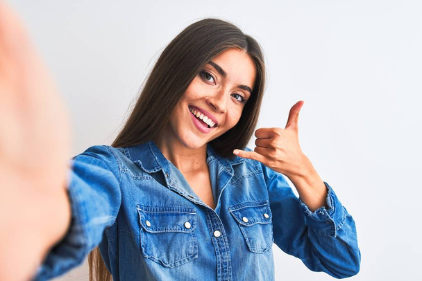 Mulher bonita vestindo camisa jeans fazer selfie por câmera sobre fundo branco isolado sorrindo fazendo gesto telefone com a mão e os dedos, como falar ao telefone. Conceitos comunicantes
. - Foto, Imagem
