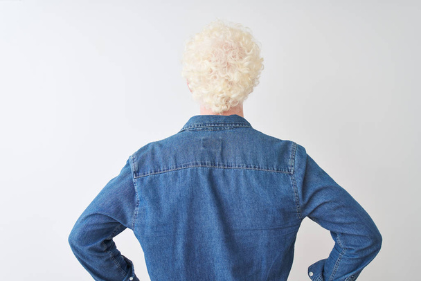 Joven hombre rubio albino con camisa de mezclilla y gafas sobre fondo blanco aislado de pie hacia atrás mirando hacia otro lado con los brazos en el cuerpo
 - Foto, imagen
