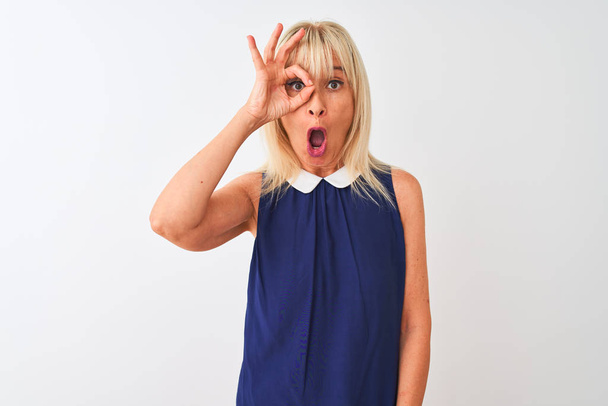 Middle age woman wearing elegant blue dress standing over isolated white background doing ok gesture shocked with surprised face, eye looking through fingers. Unbelieving expression. - Photo, Image