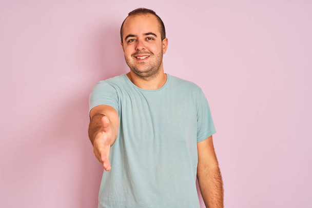Joven hombre vistiendo azul casual camiseta de pie sobre el fondo rosa aislado sonriendo amistoso ofreciendo apretón de manos como saludo y bienvenida. Negocios exitosos
. - Foto, imagen