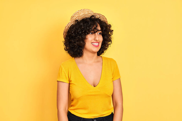 Nuori arabinainen kihara tukka yllään t-paita ja hattu yli eristetty keltainen tausta etsii pois puolelta hymy kasvoilla, luonnollinen ilme. Nauraen itsevarmana
. - Valokuva, kuva