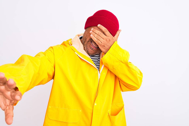 Middle age man wearing rain coat and woolen hat standing over isolated white background covering eyes with hands and doing stop gesture with sad and fear expression. Embarrassed and negative concept. - Photo, image