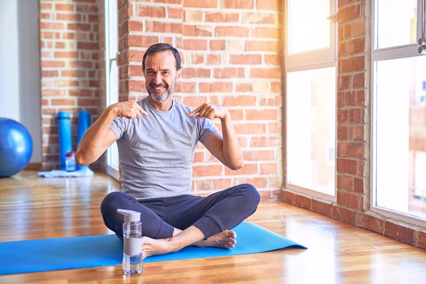 Średniego wieku przystojny sportowiec siedzi na macie robi rozciągające ćwiczenia jogi na siłowni patrząc pewnie z uśmiechem na twarzy, wskazując się palcami dumny i szczęśliwy. - Zdjęcie, obraz