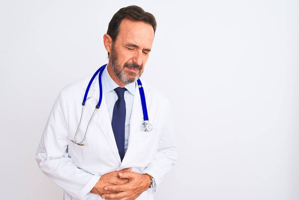 Middle age doctor man wearing coat and stethoscope standing over isolated white background with hand on stomach because indigestion, painful illness feeling unwell. Ache concept. - Foto, Imagem