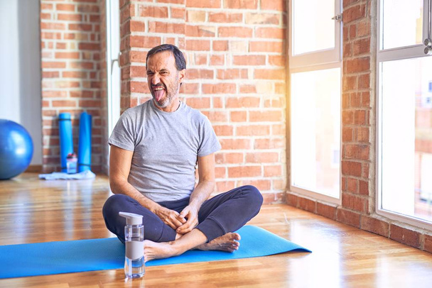 Średniego wieku przystojny sportowiec siedzi na macie robiąc rozciągające ćwiczenia jogi na siłowni wystający język szczęśliwy z zabawnym wyrazem twarzy. Koncepcja emocji. - Zdjęcie, obraz