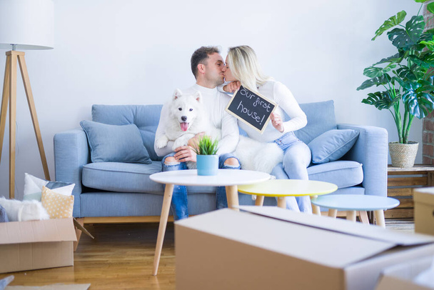 Jeune beau couple avec chien assis sur le canapé tenant tableau noir avec message embrasser à la nouvelle maison autour des boîtes en carton
 - Photo, image