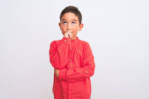 Beautiful kid boy wearing elegant red shirt standing over isolated white background with hand on chin thinking about question, pensive expression. Smiling with thoughtful face. Doubt concept. - 写真・画像