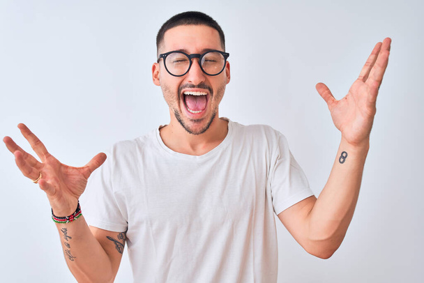 Young handsome man wearing glasses and standing over isolated background celebrating crazy and amazed for success with arms raised and open eyes screaming excited. Winner concept - Fotoğraf, Görsel