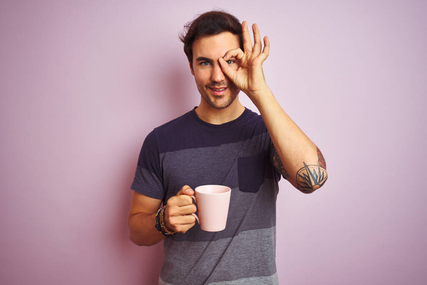 Молодий красивий чоловік з татуюванням п'є чашку кави над ізольованим рожевим тлом з щасливим обличчям, посміхаючись, роблячи знак з рукою на око, дивлячись через пальці
 - Фото, зображення