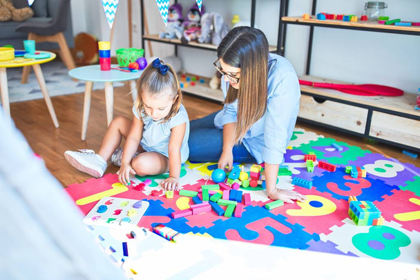 Valkoihoinen tyttö leikkii ja oppii leikkikoulussa naisopettajan kanssa. Äiti ja tytär leikkihuoneessa lelujen ympärillä leikkimässä pullistumilla
 - Valokuva, kuva