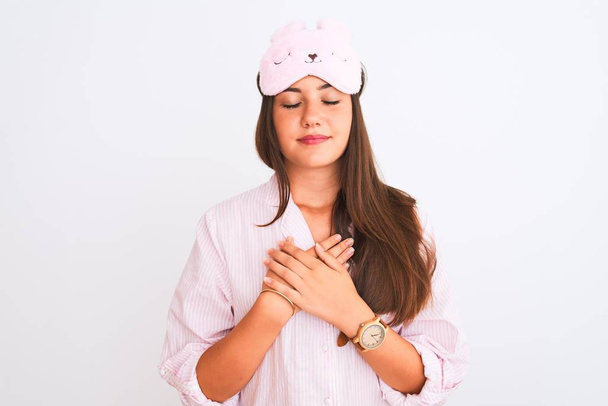 Young beautiful girl wearing pajama and sleep mask standing over isolated white background smiling with hands on chest with closed eyes and grateful gesture on face. Health concept. - Photo, Image