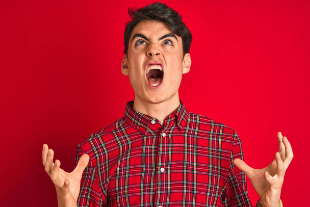 Teenager boy wearing red shirt standing over isolated background crazy and mad shouting and yelling with aggressive expression and arms raised. Frustration concept. - Fotoğraf, Görsel