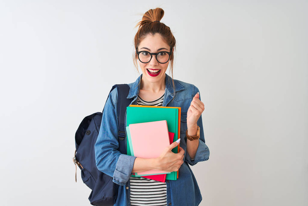 Redhead student woman wearing backpack holding books over isolated white background screaming proud and celebrating victory and success very excited, cheering emotion - Photo, image