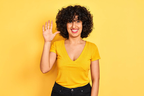 Νεαρή αραβίδα γυναίκα με σγουρά μαλλιά φορώντας t-shirt στέκεται πάνω από απομονωμένο κίτρινο φόντο δείχνει και δείχνει προς τα πάνω με τα δάχτυλα αριθμό πέντε, ενώ χαμογελά αυτοπεποίθηση και χαρούμενος. - Φωτογραφία, εικόνα
