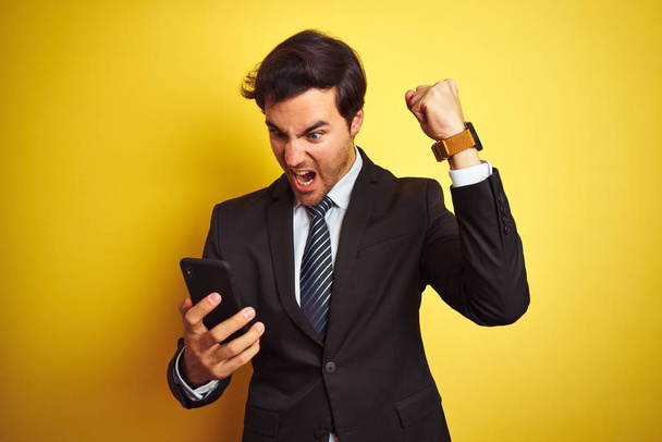Young handsome businessman using smartphone standing over isolated yellow background annoyed and frustrated shouting with anger, crazy and yelling with raised hand, anger concept - Foto, Bild