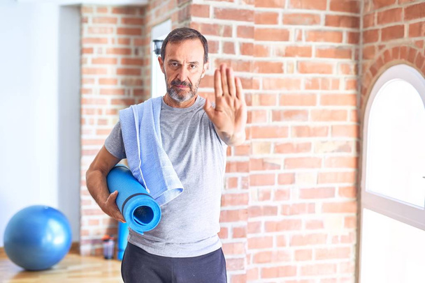 Middle age handsome sportman holding mat and towel standing before do exercise at gym with open hand doing stop sign with serious and confident expression, defense gesture - Photo, Image
