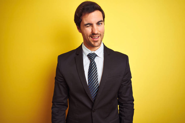 Joven hombre de negocios guapo con traje y corbata de pie sobre fondo amarillo aislado guiño mirando a la cámara con expresión sexy, cara alegre y feliz
. - Foto, Imagen