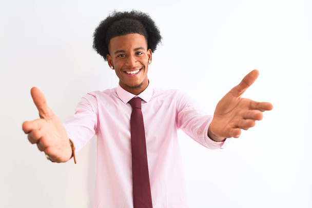 Mladý Afričan americký obchodník na sobě nosí kravatu stojící nad izolovaným bílým pozadím a dívá se na kameru usmívající se otevřenou náručí pro objetí. Veselý výraz zahrnující štěstí. - Fotografie, Obrázek