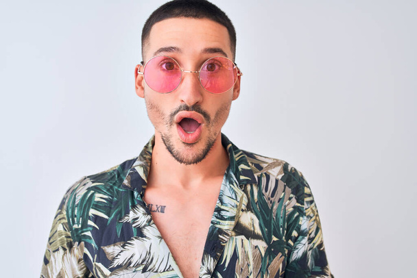Jonge knappe man draagt Hawaïaanse sumer shirt en roze zonnebril over geïsoleerde achtergrond bang in shock met een verrassing gezicht, bang en opgewonden met angst uitdrukking - Foto, afbeelding