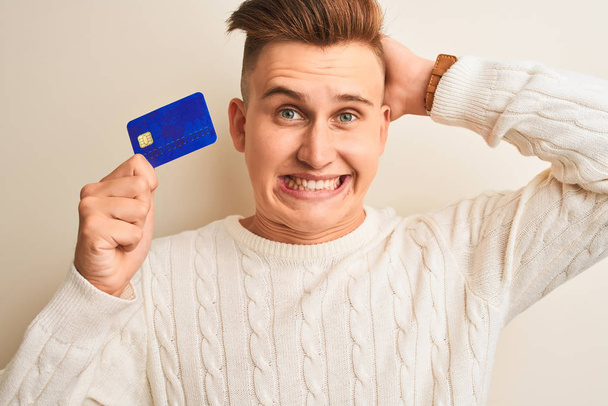 Mladý pohledný muž držící kreditní kartu nad izolovaným bílým pozadím, s rukou na hlavě, šokovaný hanbou a překvapením, rozzlobený a frustrovaný. Strach a zmatek z omylu. - Fotografie, Obrázek