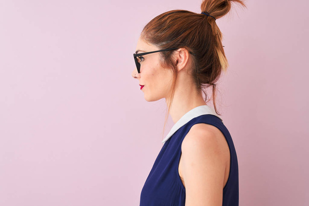 Piros hajú nő copfos visel elegáns ruha és szemüveg felett elszigetelt rózsaszín háttér keres oldalra, pihenjen profil pózol természetes arc magabiztos mosollyal. - Fotó, kép