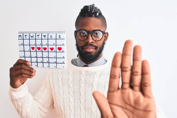 Afrykański Amerykanin z warkoczami trzymając kalendarz okresu na odizolowanym białym tle z otwartą ręką robi znak stop z poważnym i pewnym siebie wyrazem, gest obrony - Zdjęcie, obraz
