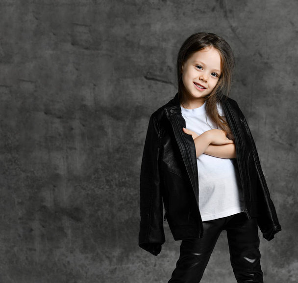 Small smiling cute girl in black and white rock star style casual clothing and white sneakers standing over grey concrete background - Photo, image