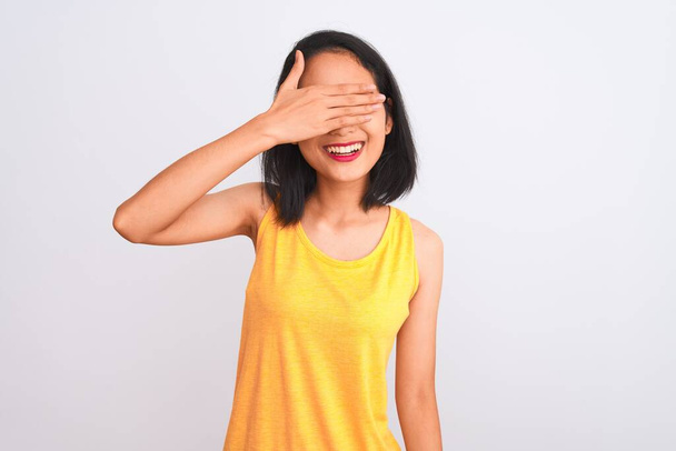 Mujer china joven con camiseta casual amarilla de pie sobre fondo blanco aislado sonriendo y riendo con la mano en la cara cubriendo los ojos para sorpresa. Concepto ciego
. - Foto, Imagen