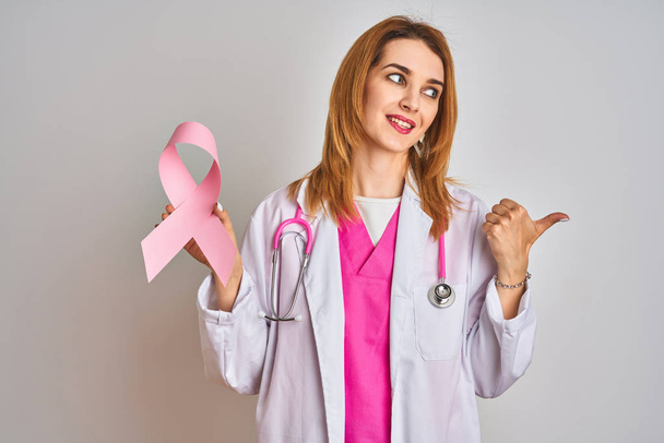 Rossa donna medico caucasico che tiene il nastro rosa cancro su sfondo isolato puntando e mostrando con il pollice verso l'alto di lato con il viso felice sorridente
 - Foto, immagini