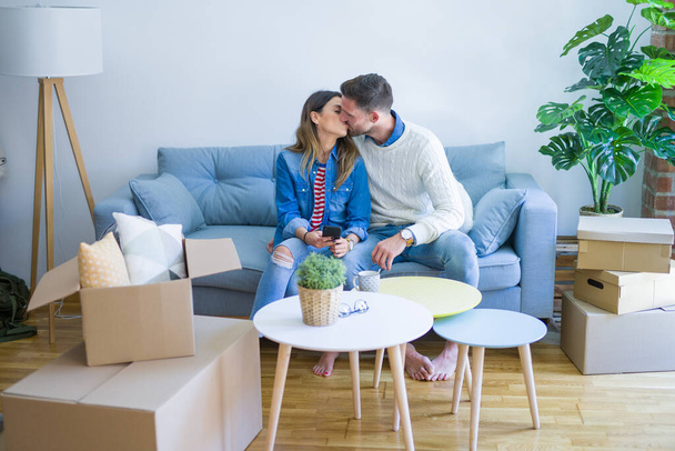 Young beautiful couple sitting on the sofa drinking cup of coffee using smartphone kissing at new home around cardboard boxes - Photo, Image