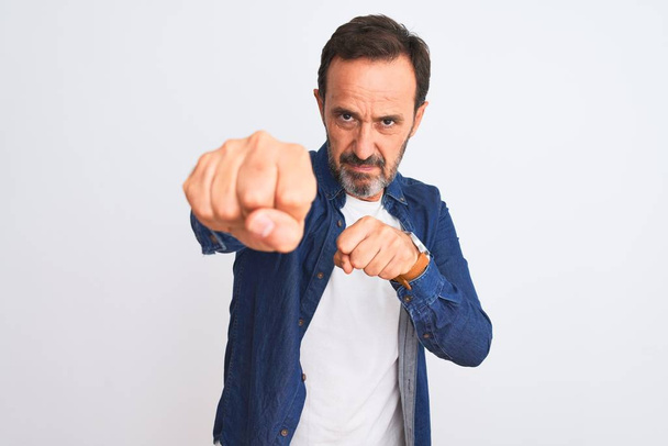 Middle age handsome man wearing blue denim shirt standing over isolated white background Punching fist to fight, aggressive and angry attack, threat and violence - Photo, Image
