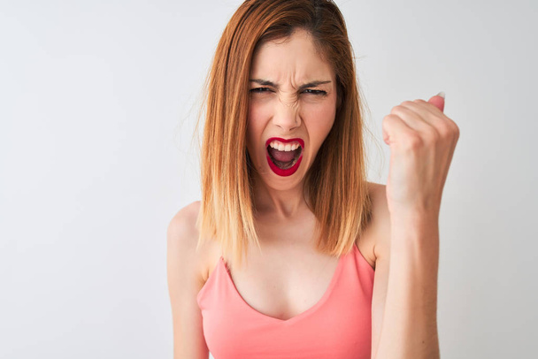 Beautiful redhead woman wearing casual pink t-shirt standing over isolated white background annoyed and frustrated shouting with anger, crazy and yelling with raised hand, anger concept - Fotó, kép