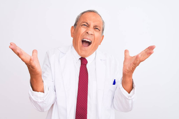 Senior grey-haired scientist man wearing coat standing over isolated white background crazy and mad shouting and yelling with aggressive expression and arms raised. Frustration concept. - Photo, Image