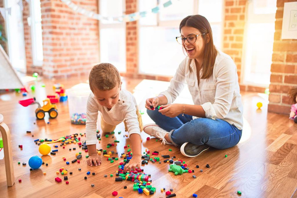 Воспитатель и малыш играют со строительными блоками вокруг множества игрушек одновременно.
 - Фото, изображение