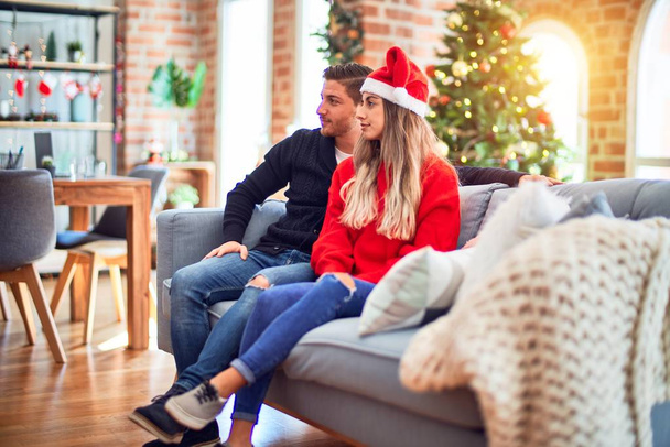Fiatal pár visel télapó claus kalap ül a kanapén körül karácsonyfa otthon keres oldalra, pihenjen profil pózol természetes arc magabiztos mosollyal. - Fotó, kép