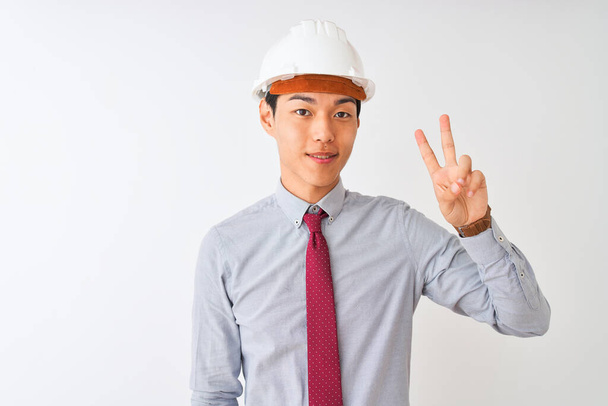 Κινέζος αρχιτέκτονας άνδρας φορώντας γραβάτα και κράνος στέκεται πάνω από απομονωμένο λευκό φόντο δείχνει και δείχνει προς τα πάνω με τα δάχτυλα νούμερο δύο, ενώ χαμογελά αυτοπεποίθηση και χαρούμενος. - Φωτογραφία, εικόνα