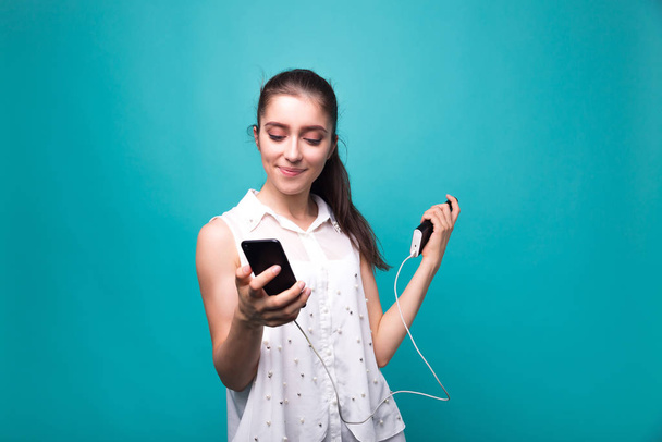 Девушка с телефоном и банк власти в руках Молодая красивая женщина улыбается с телефоном и зарядки в его руке синий голубой фон студии
 - Фото, изображение