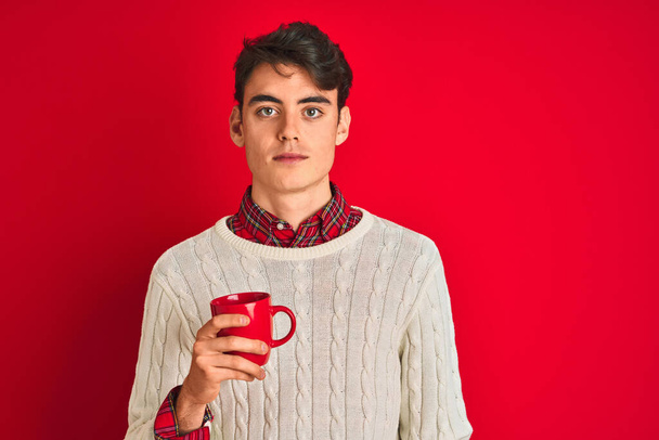 Adolescente niño usando suéter de invierno bebiendo una taza de café sobre fondo rojo aislado con una expresión de confianza en la cara inteligente pensando en serio
 - Foto, imagen