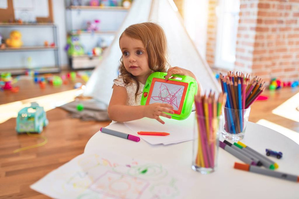 Чарівна блондинка малюк малює, використовуючи цифрову дошку навколо великої кількості іграшок в дитячому саду
 - Фото, зображення