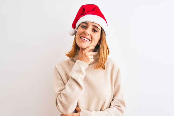 Красива руда жінка в різдвяному капелюсі над ізольованим тлом впевнено дивиться на камеру, посміхаючись схрещеними руками і піднятою рукою на підборідді. Мислення позитивне
. - Фото, зображення